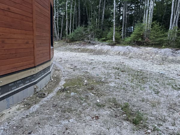 The base of the building from outside: wood siding above, the crawlspace wall wrapped in insulation board, a gravel apron and level ground running into the trees