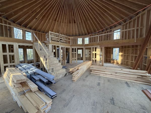Inside: the rafter fan overhead, interior stud walls framed, the stair to the loft built, and bundles of flooring, cladding and metal roof panels stacked on the subfloor