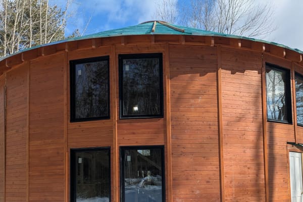 The wood-clad shell with black-framed windows in snow, the roof deck still under green membrane
