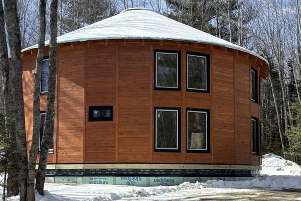 Snow on the roof membrane, the wood shell, the insulated crawlspace band at the base and level ground with birch and spruce behind