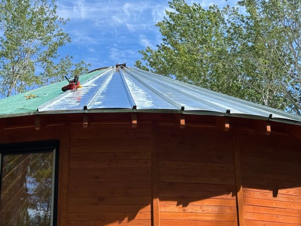 Standing seam metal panels half installed over the green membrane, trees and blue sky behind