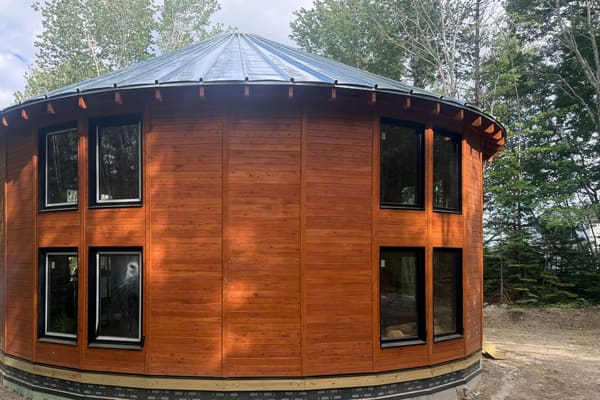 The shell finished: the standing seam roof cone, wood walls, black windows and the crawlspace band on a flat gravel pad in summer woods