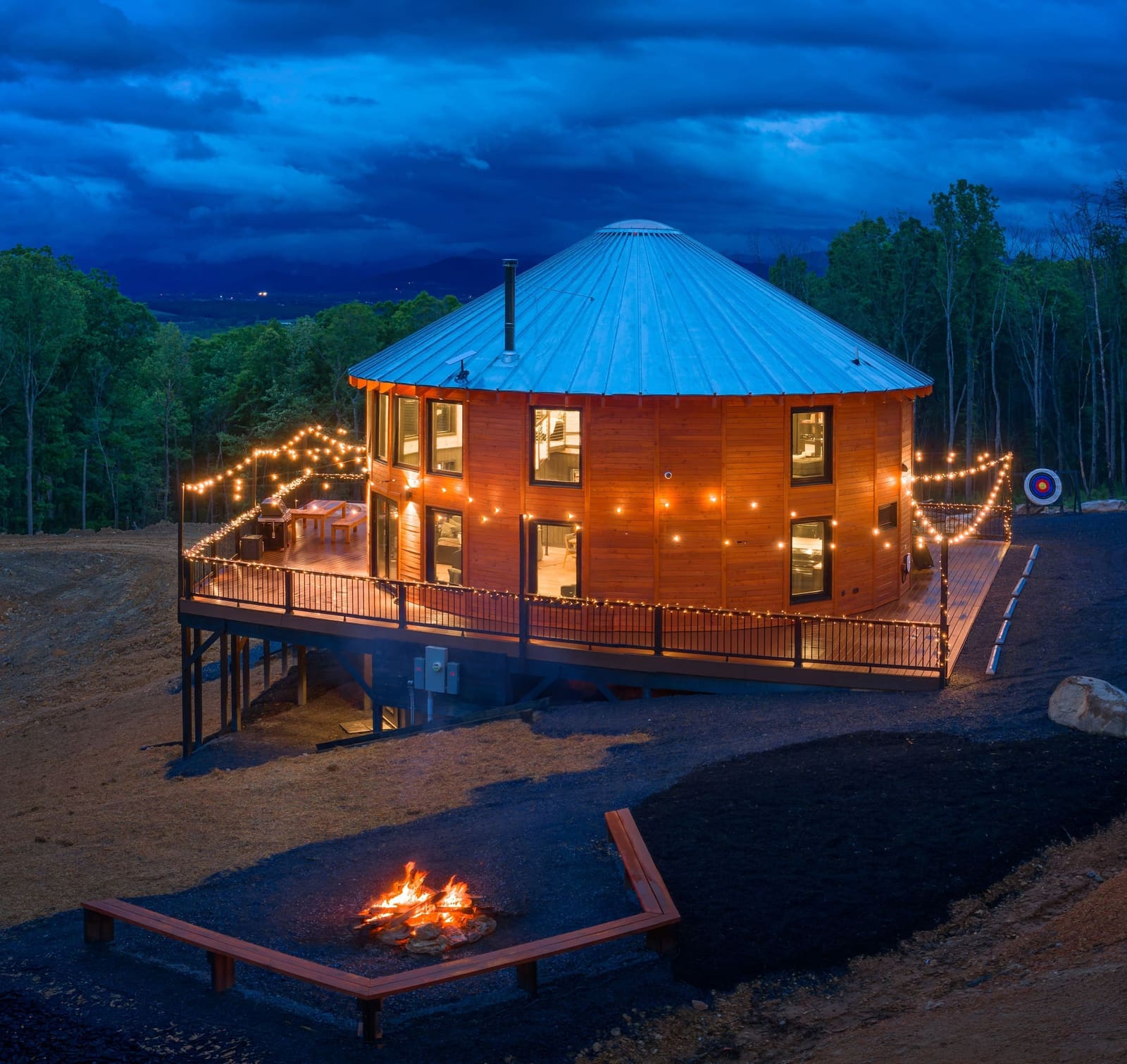 Luray Yurt at twilight: a round wood building with a metal cone roof, lit windows, a deck with string lights and a fire pit in front