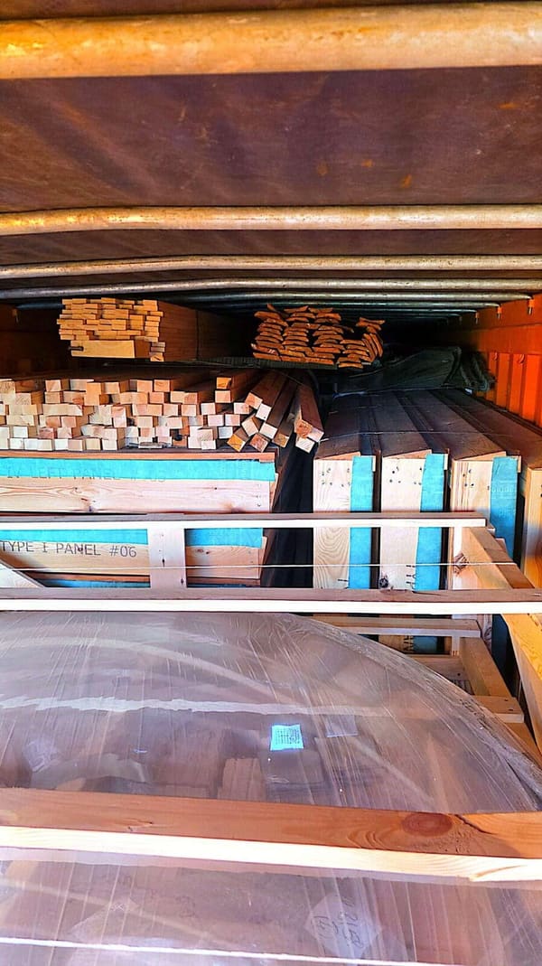 Inside the container, numbered panel crates with blue label bands stacked above the wrapped acrylic dome, rafter bundles on top
