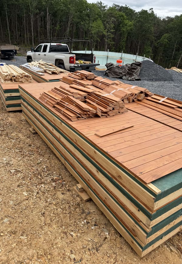 Stacked wall panel crates with green edge protection, decking and cladding boards bundled on top, a pickup and the basement wall behind