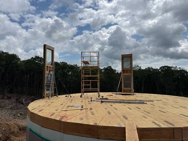 The round plywood deck over the basement with three tall glazed panels standing braced, a ladder and a rolling scaffold