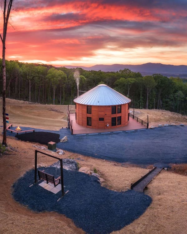 The building at sunset from above: the metal roof, larch siding, the deck, a gravel drive, a fire pit and the mountains behind
