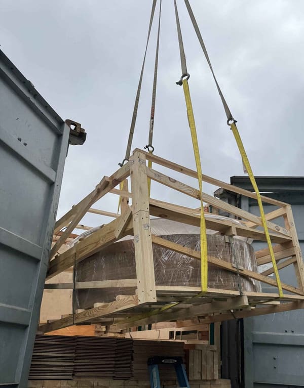 A crane lifting a strapped timber crate up out of the open-top container, the container doors open