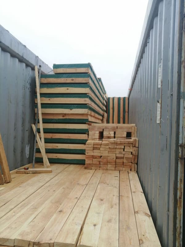 Inside the open-top container, stacked panels with green edge protection and bundled lumber on a timber floor