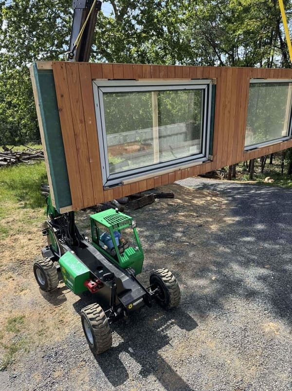 A green telehandler carrying a clad wall panel with a large window across the site